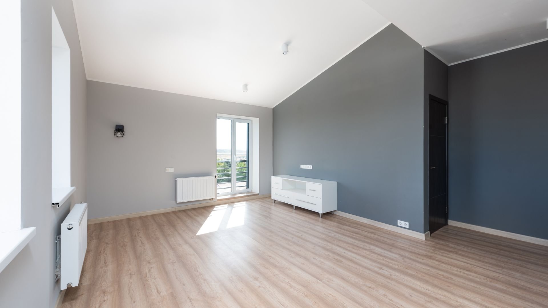 Empty room with gray walls, wooden floor, white furniture, and large window